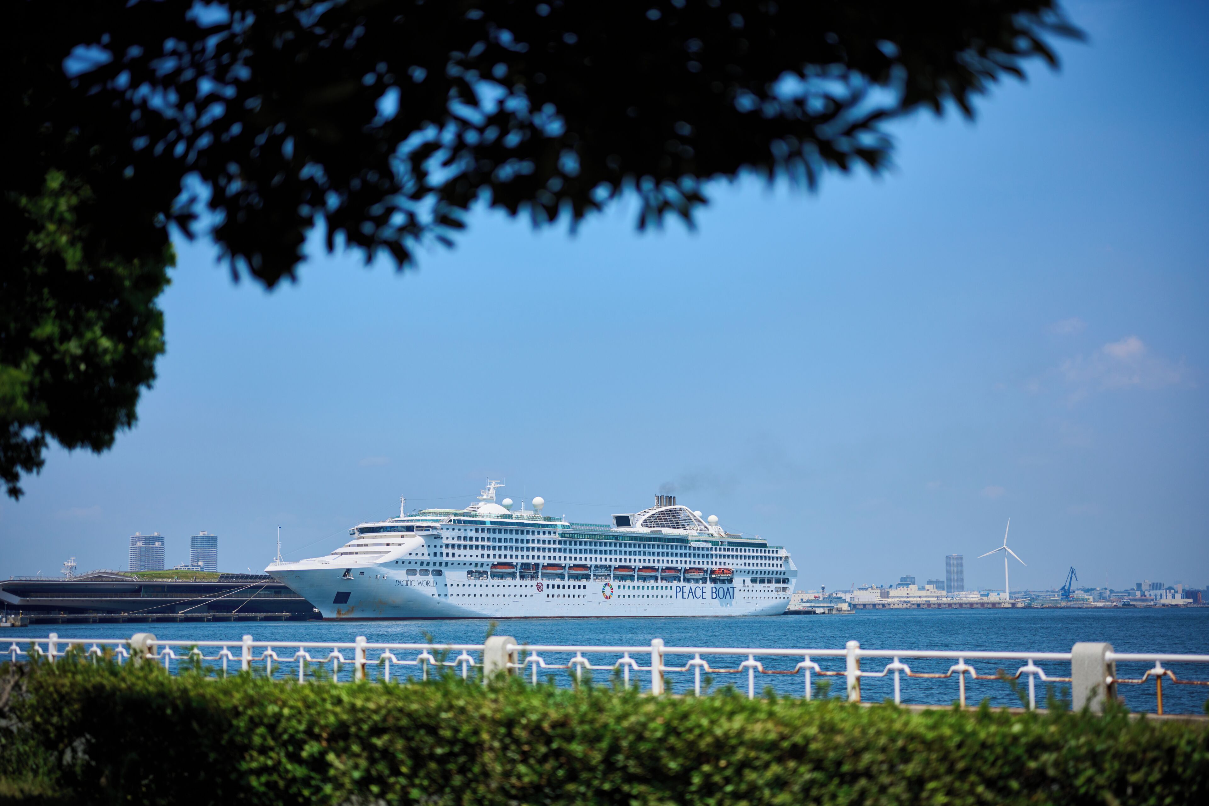 Un gran crucero amarrado en un puerto, con un horizonte urbano y aerogeneradores al fondo.