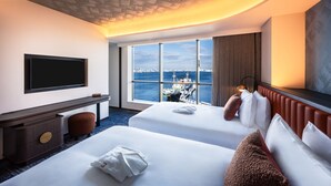 Ein Hotelzimmer mit zwei Betten, einem Fernseher, einem Schreibtisch und einem großen Fenster mit Blick auf die Stadt.