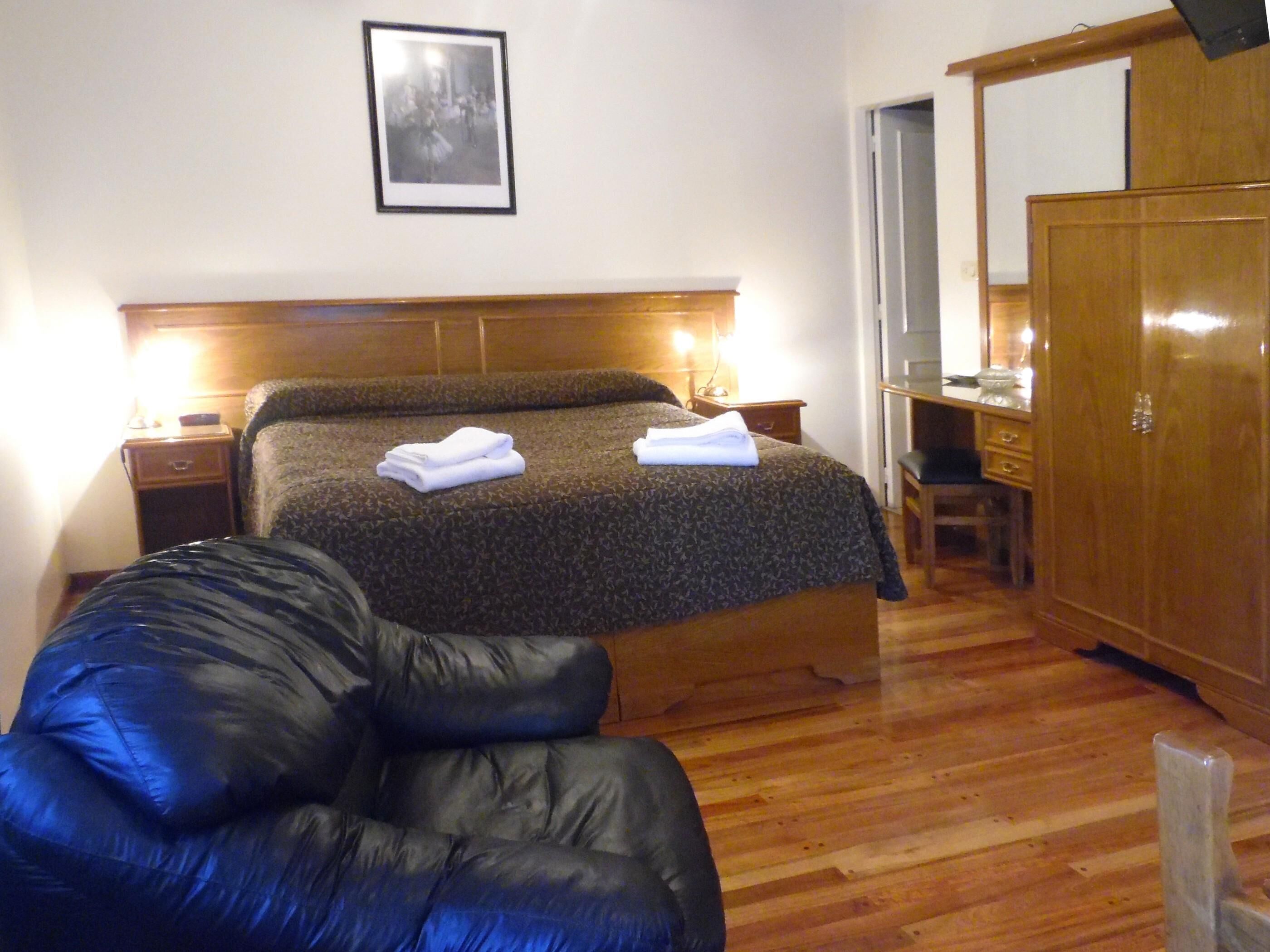 A bedroom with a bed, a wooden wardrobe, a desk, and a chair.