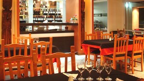 A restaurant with wooden chairs and tables, a bar area with wine bottles, and a view of the kitchen through a window.