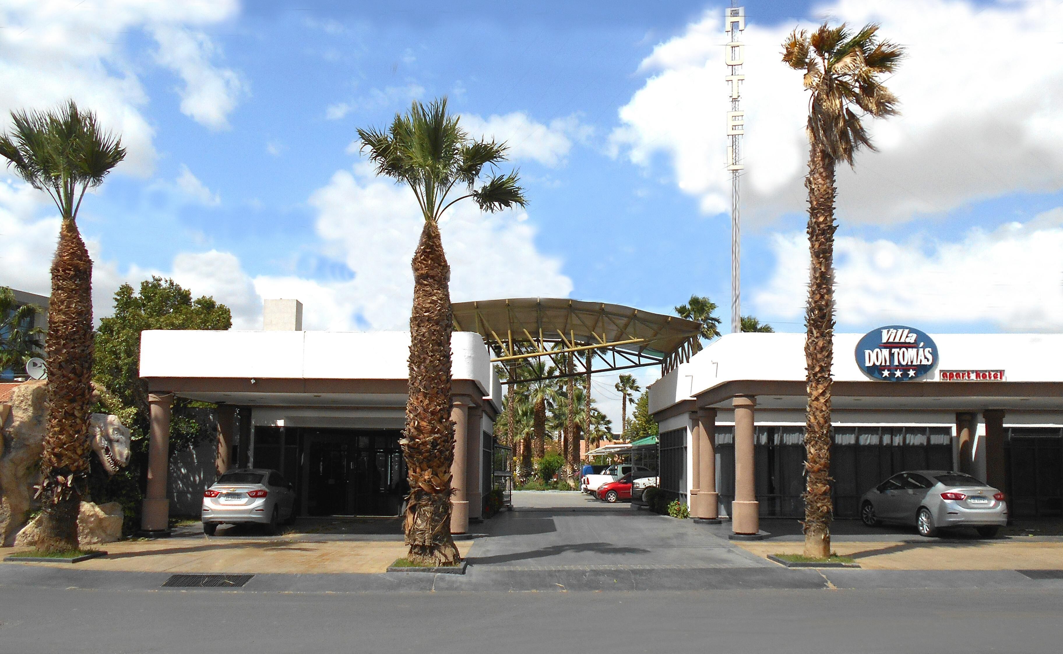 A hotel entrance with palm trees, a covered walkway, and parked cars.