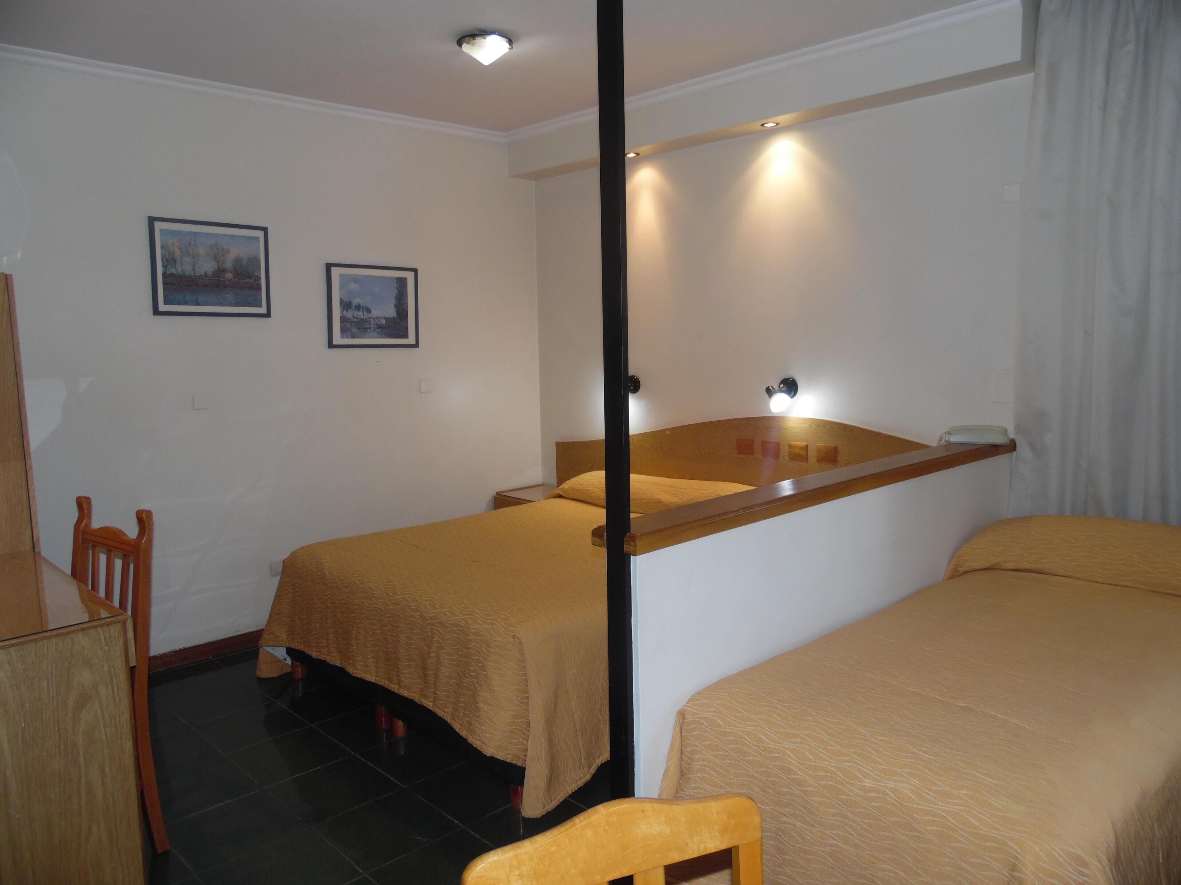A hotel room with two single beds, a wooden chair, a desk, and two framed pictures on the wall.