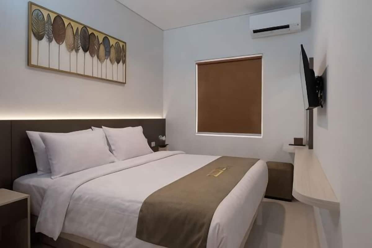 A hotel room with a bed, a wall-mounted TV, an air conditioning unit, and a window with blinds.