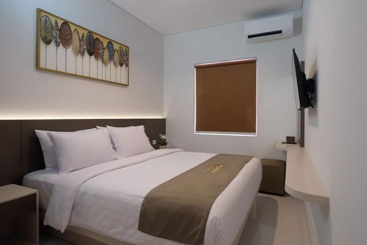 A neatly arranged hotel room with a bed, a bedside table, a wall-mounted TV, and a window with blinds.