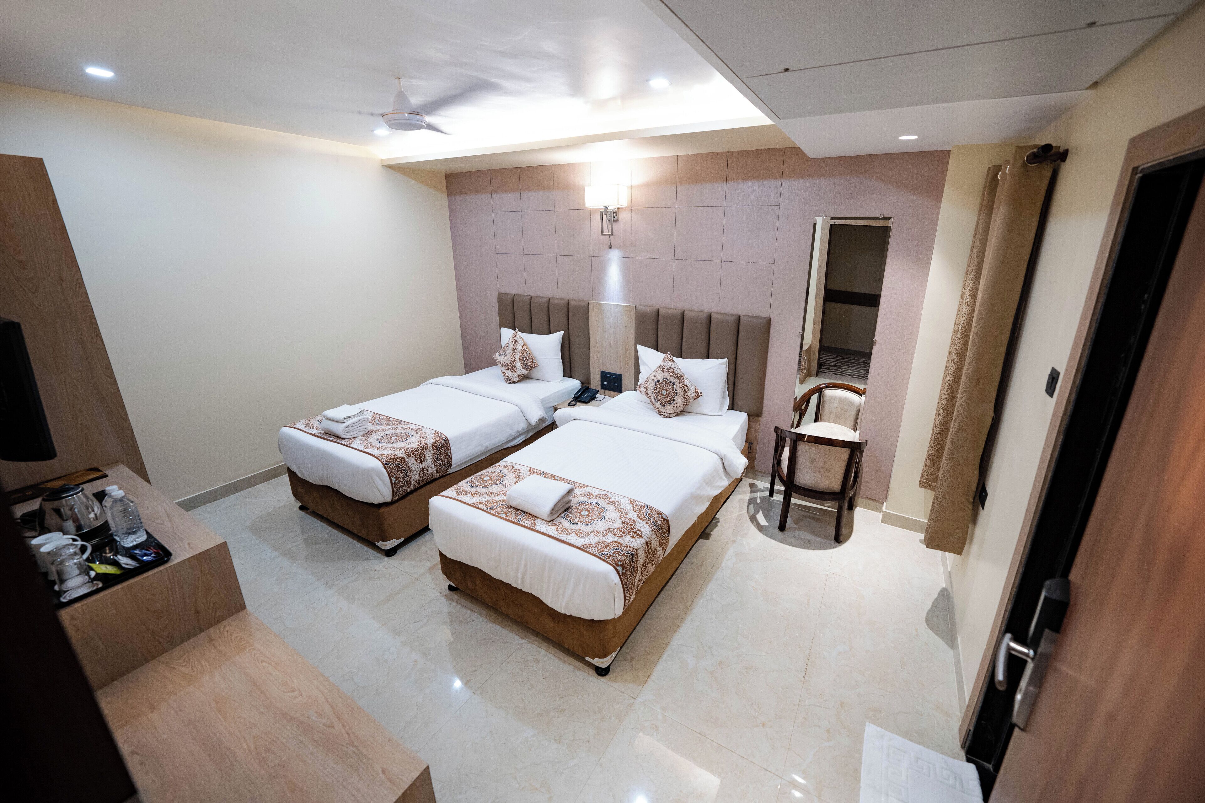 A hotel room with two beds, a desk with a chair, a small table, and a bathroom with a mirror and towel rack.