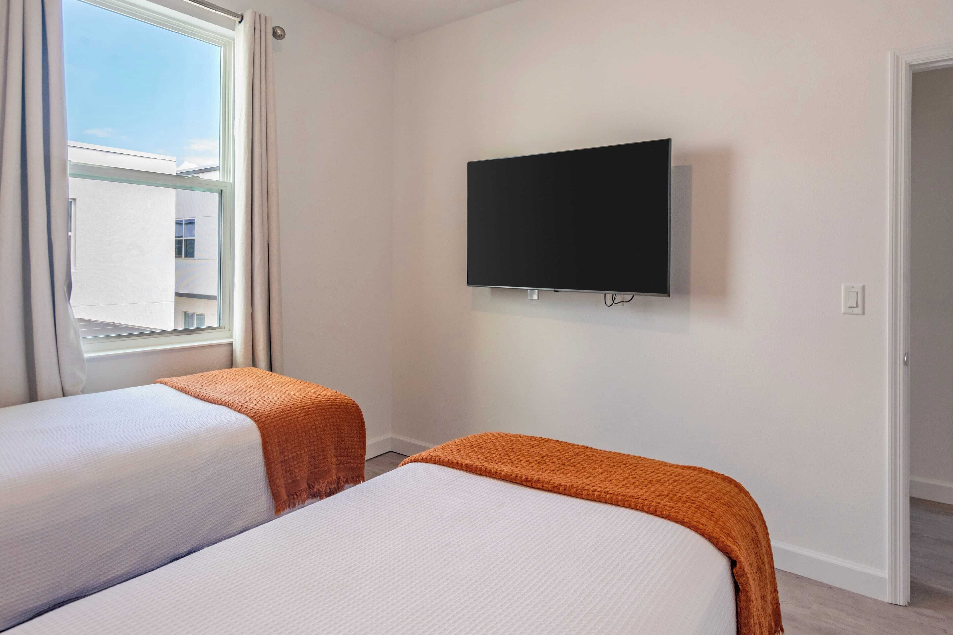 Une chambre d’hôtel avec deux lits, un téléviseur fixé au mur et une fenêtre avec des rideaux.