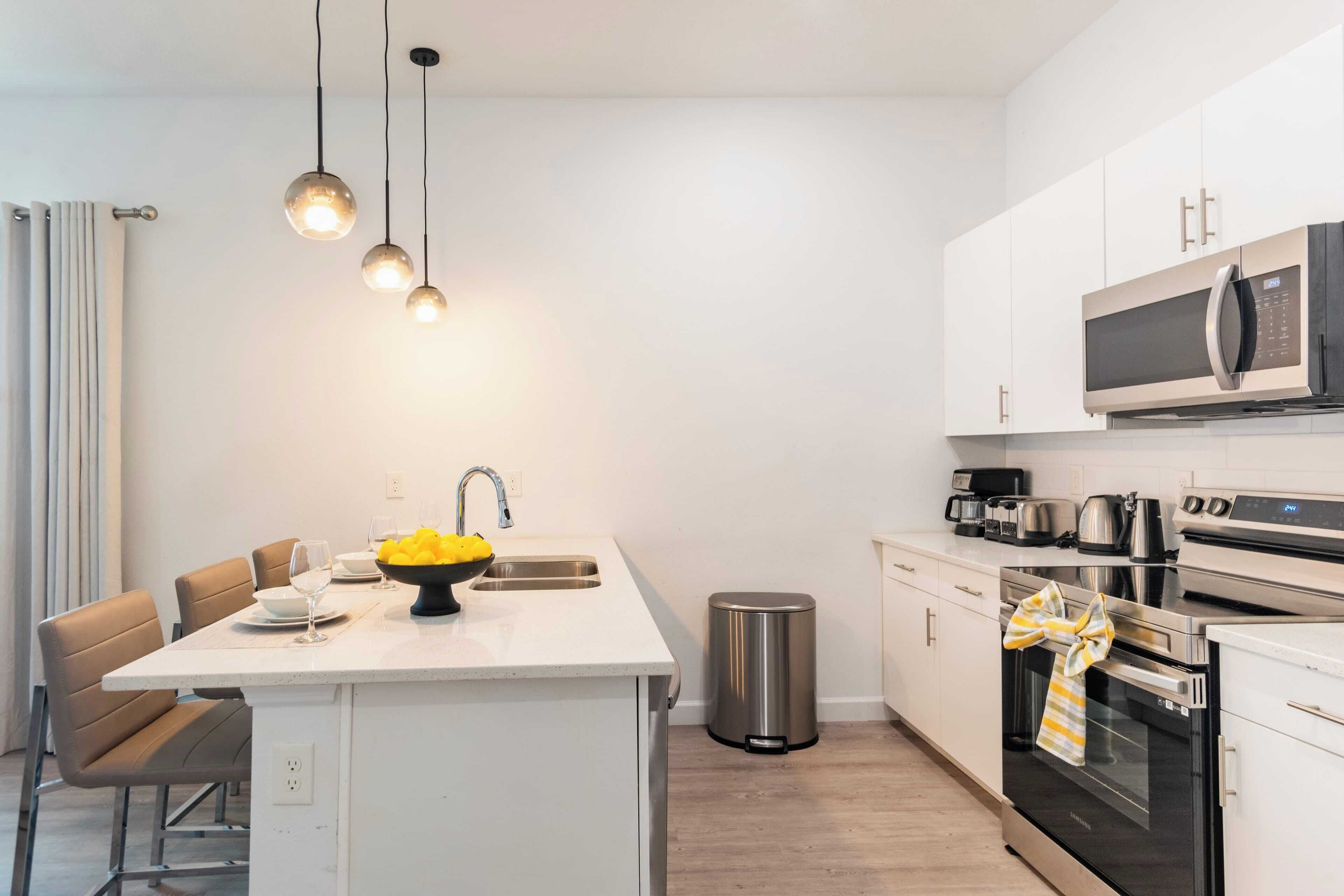 Une cuisine moderne avec des meubles blancs, des appareils en acier inoxydable, un îlot central agrémenté d’un panier de fruits et des luminaires suspendus.