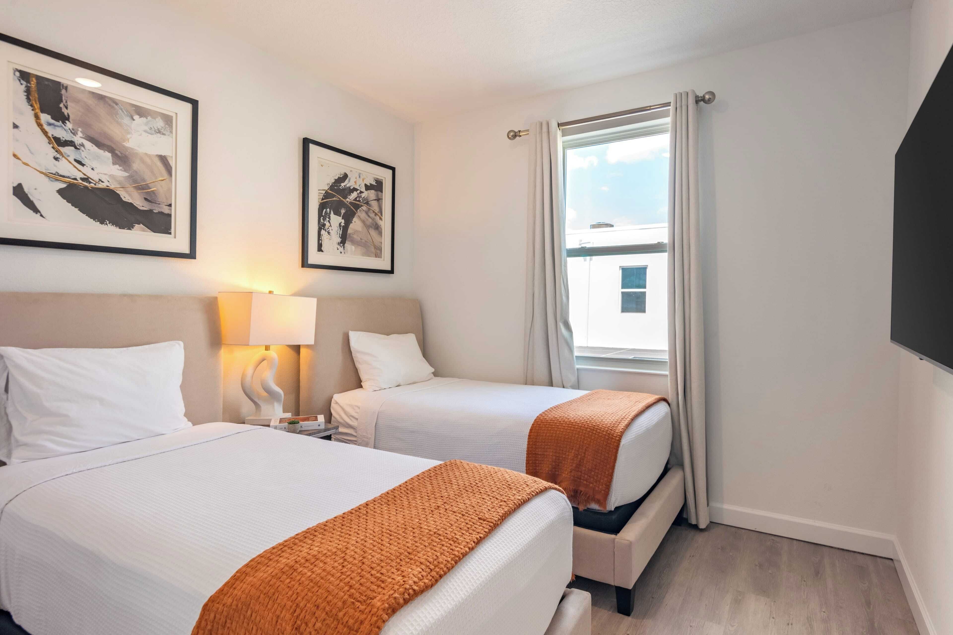 A hotel room with two beds, a window with curtains, a wall-mounted TV, and framed artwork.