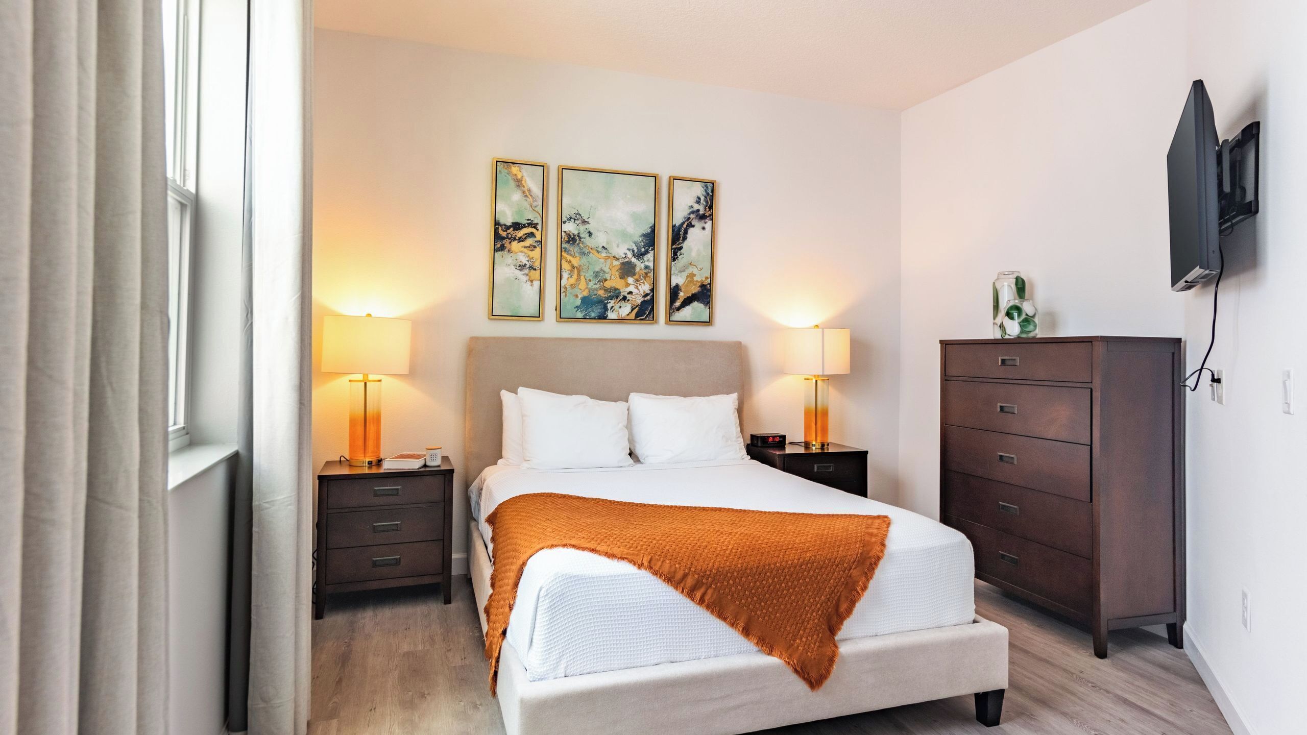 A hotel room with a bed, two bedside tables, a dresser, a TV mounted on the wall, and three abstract paintings above the bed.