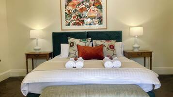 Una cama bien hecha con sábanas blancas, dos almohadas y un cojín decorativo con motivos florales. Hay dos mesitas de noche con lámparas, un banco tapizado en verde y una obra de arte enmarcada sobre la cama.