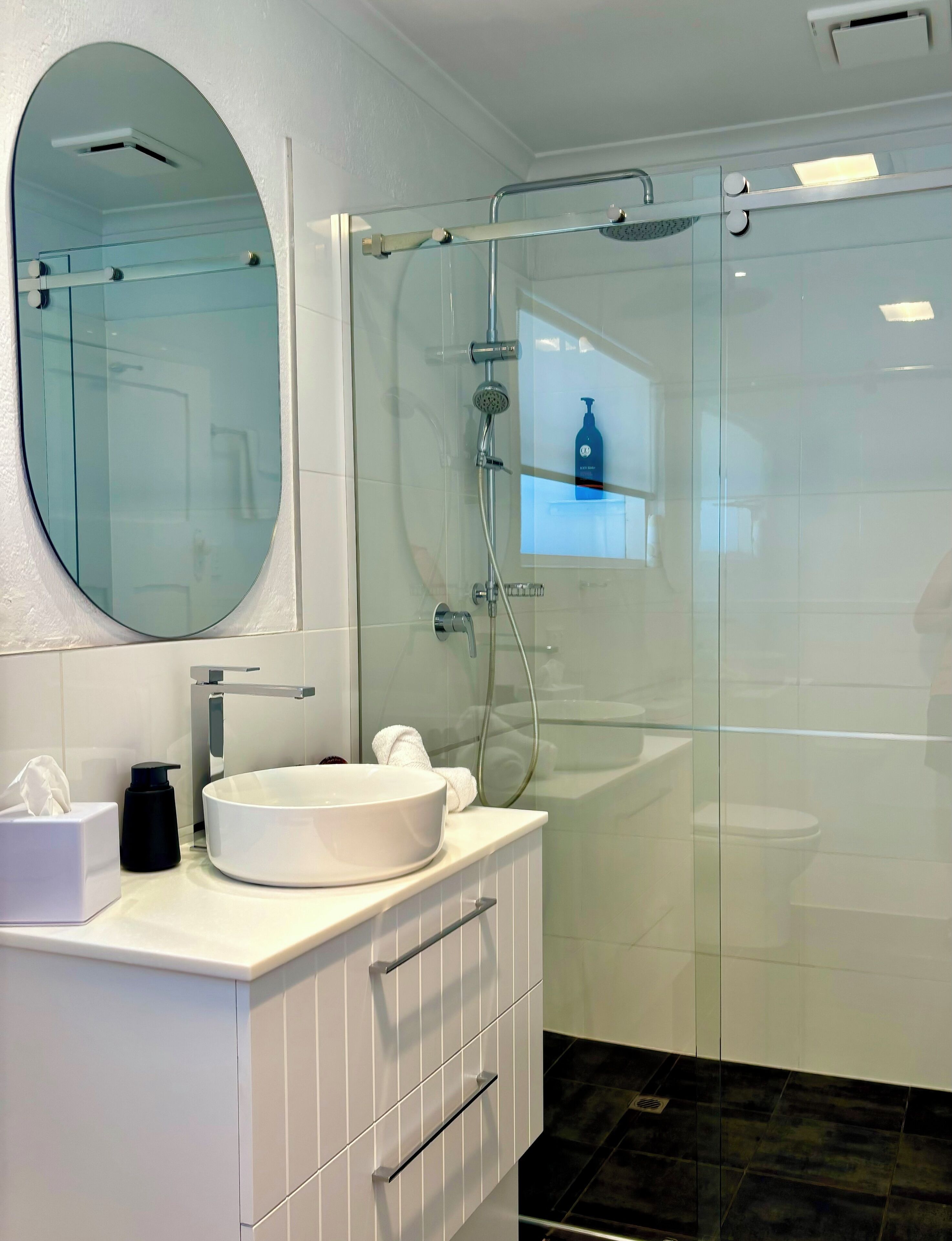 A modern bathroom with a round mirror, a white sink, a glass-enclosed shower, and a small window above the sink.