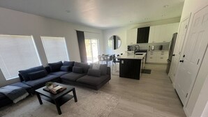 Uma sala de estar moderna com um sofá modular, uma mesa de centro e um espaço de refeições, com uma cozinha ao fundo.