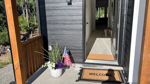 Um deck de madeira com um tapete de boas-vindas e um vaso de plantas com flores e uma bandeira americana.