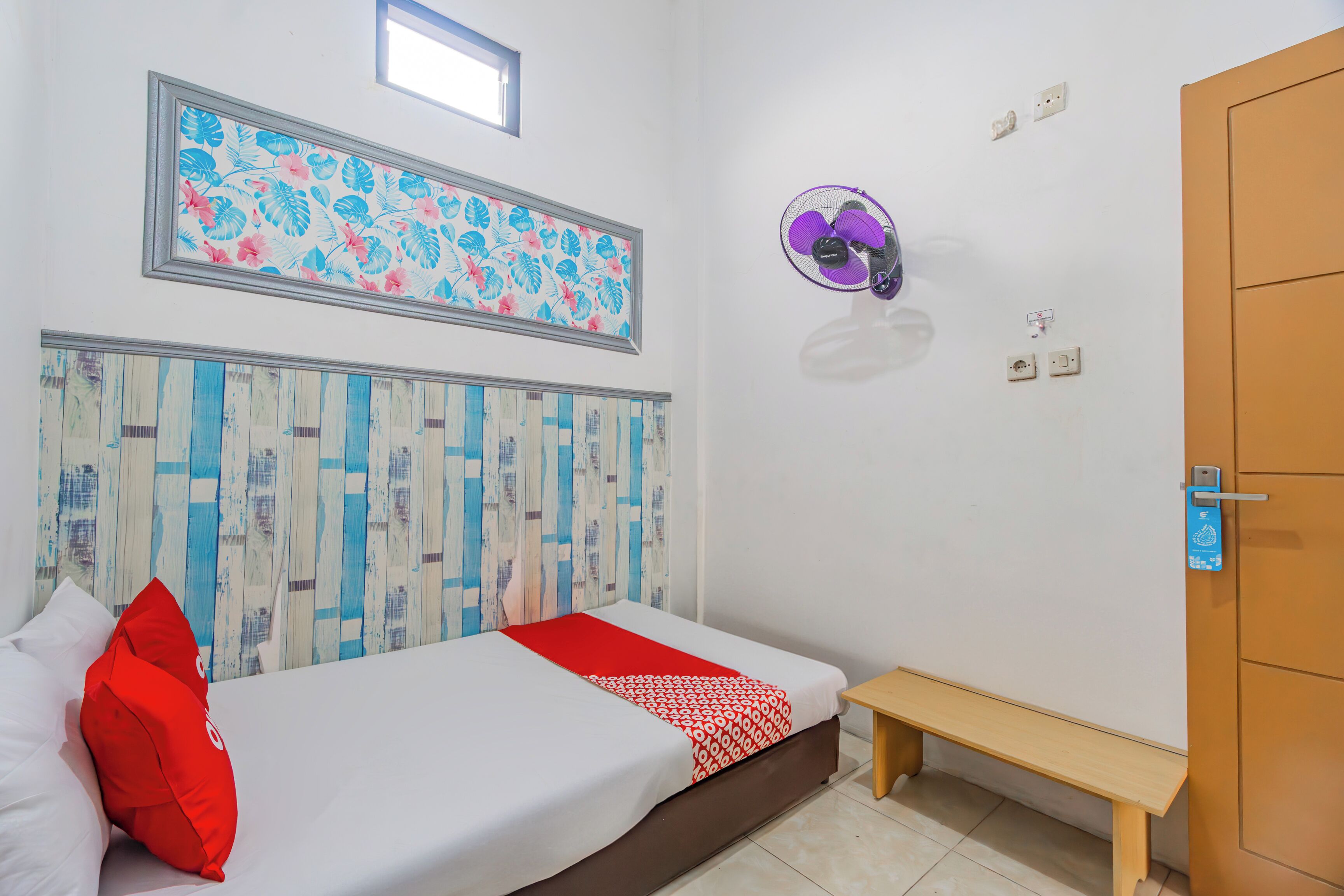 A bedroom with a bed, a bench, a wall-mounted fan, and a door.