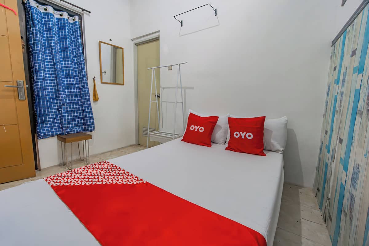 A bedroom with a bed, two red pillows with "OYO" text, a blue checkered curtain, a wooden door, and a mirror on the wall.