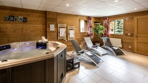 A spa room with a hot tub, lounge chairs, wooden walls, and a window with a view of trees.