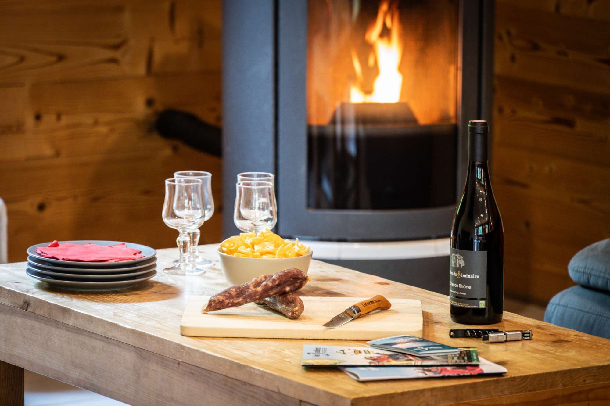 A wooden table with plates, glasses, a bowl of snacks, a bottle of wine, a knife, and magazines. A fireplace with a fire is in the background.