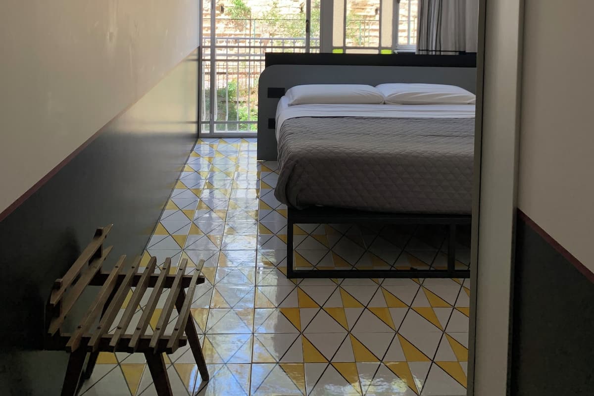 A room with a bed, a bench, and a view through a window with a metal grid.