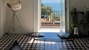 Ein Zimmer mit Schachbrettmuster-Boden, einem Tisch mit Büchern, einer Stehlampe und Meerblick vom Balkon.
