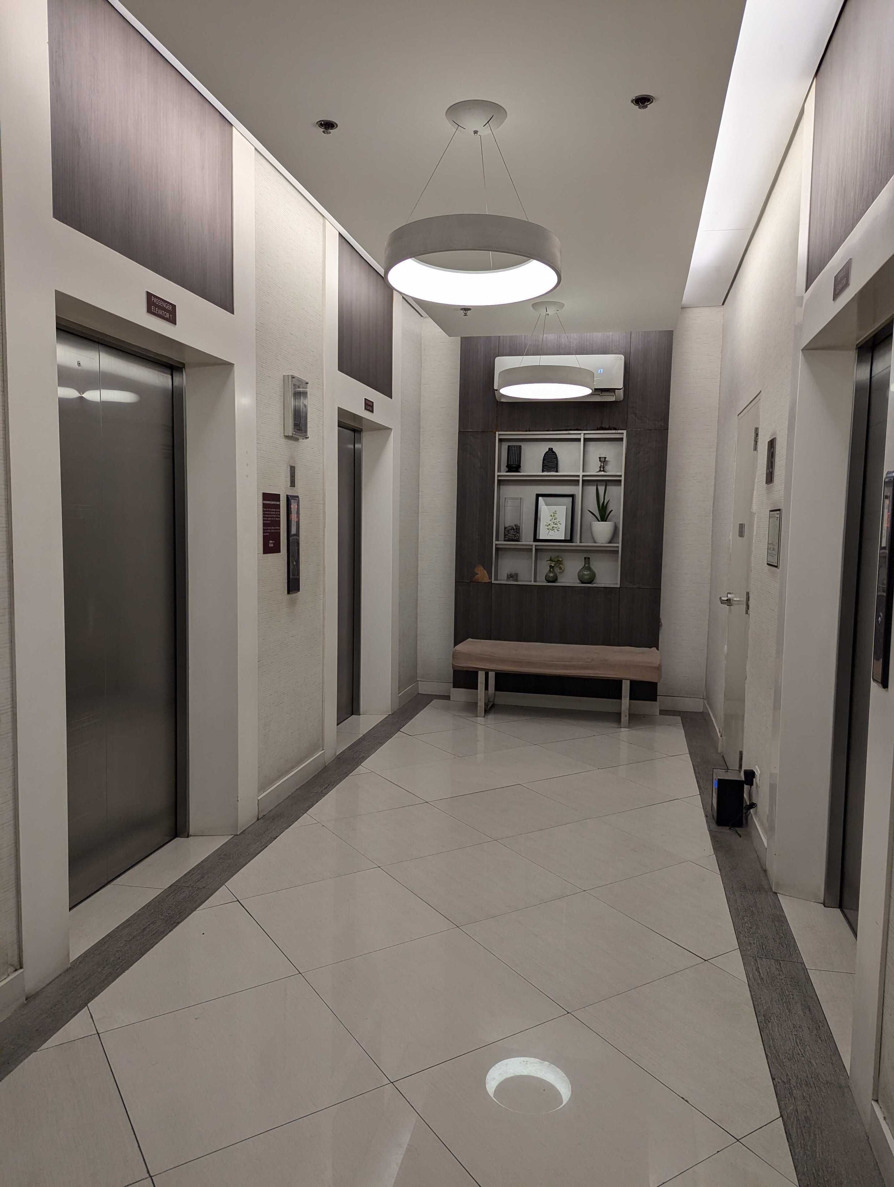 A modern elevator area with a bench, a wall-mounted shelf with decorative items, and a ceiling light fixture.