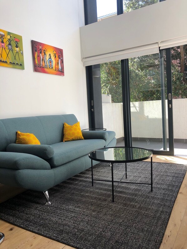 A modern living room with a blue sofa, two yellow pillows, a glass coffee table, and a black rug.