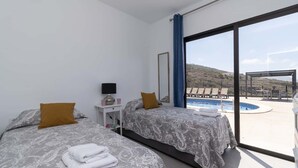 A modern hotel room with two beds, a large window with a view of a pool and outdoor seating, and a small table with a lamp.