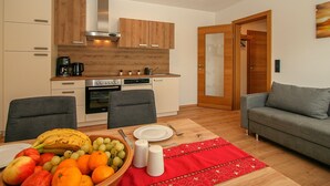 Ein gemütlicher Wohnraum mit Küchenzeile, einem Essbereich mit Obst und einer roten Tischmatte sowie einem grauen Sofa.