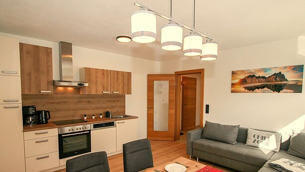 Eine moderne Küche mit Holzausstattung, ein Essbereich mit Tisch für zwei Personen und ein graues Sofa im Wohnzimmer.