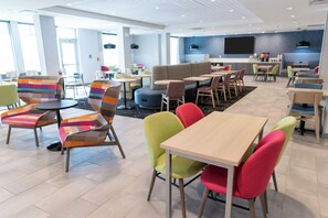 A modern hotel lobby with seating areas, a kitchen, and a large TV screen.