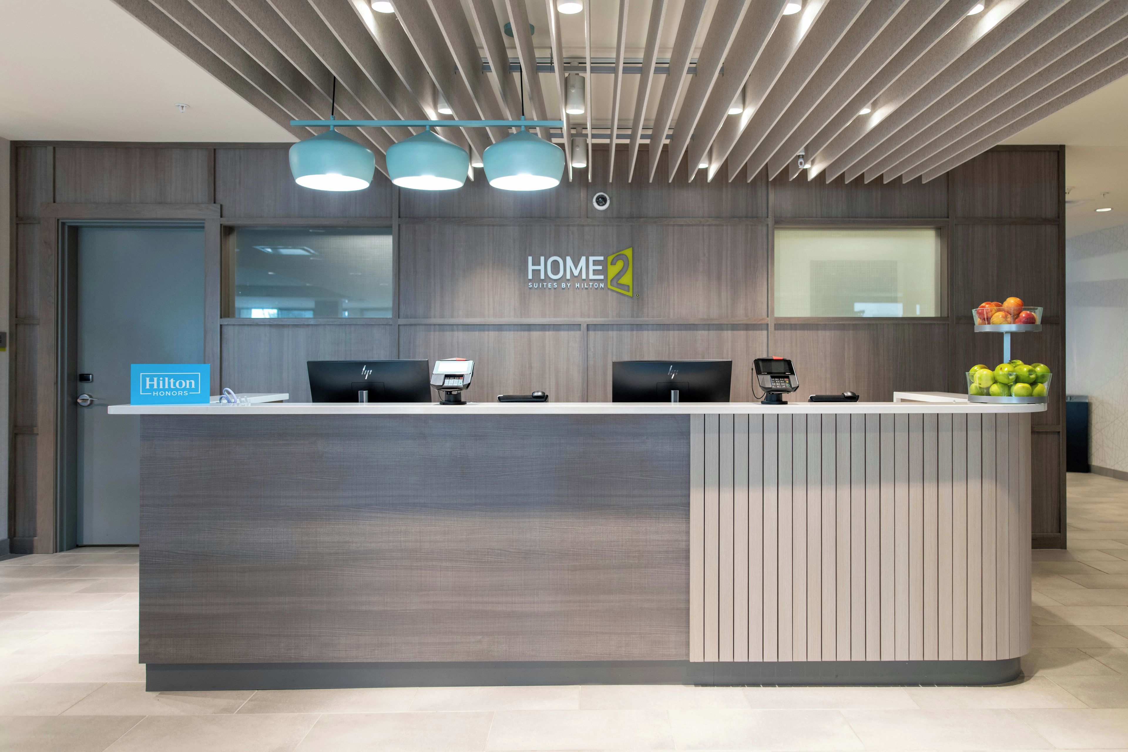 Una moderna reception hotel con un cartello 'HOME 2', due monitor computer, un telefono e un espositore di frutta.