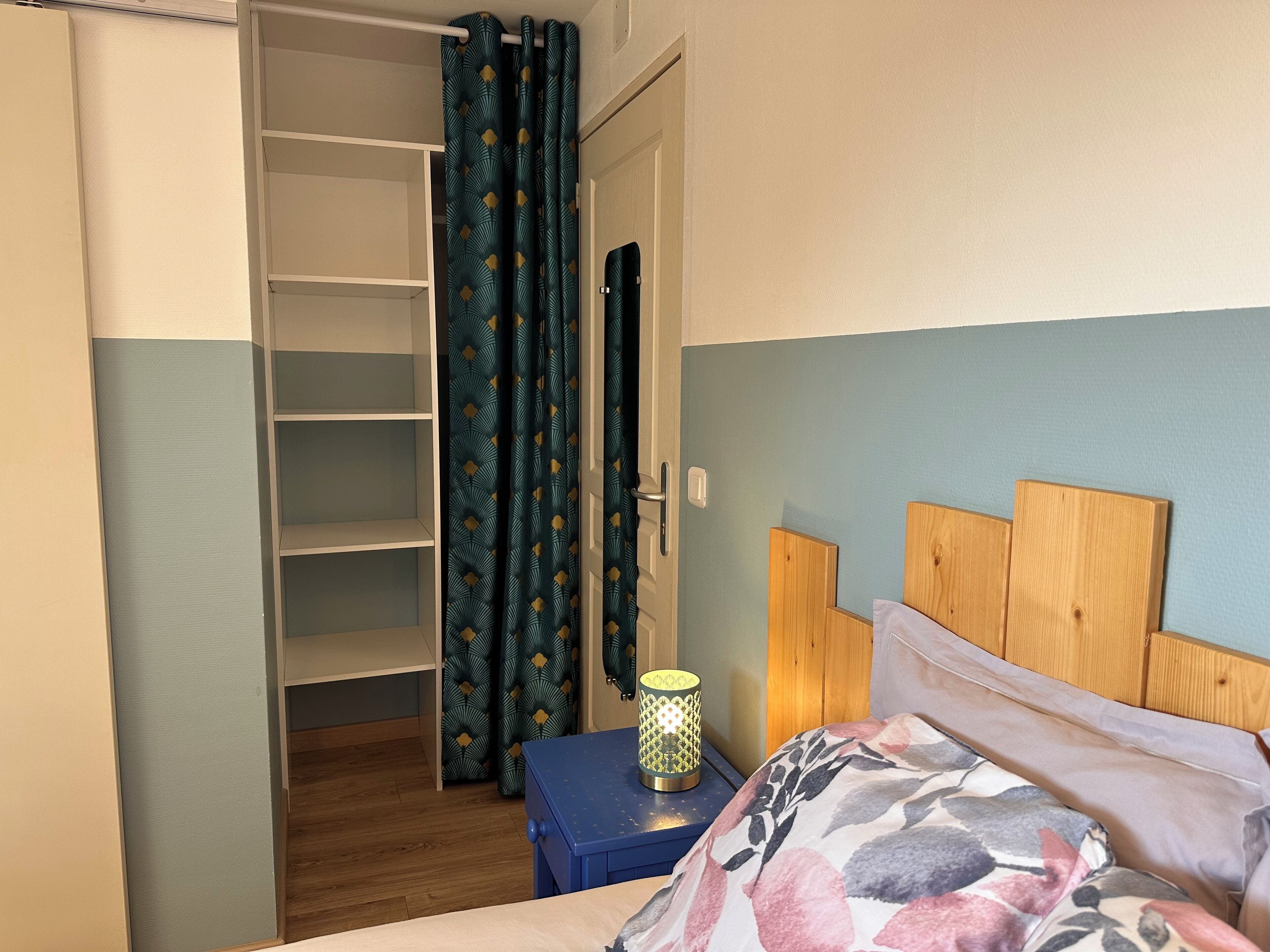 A bedroom with a bed, a nightstand, a lamp, a wardrobe, and a door with curtains.