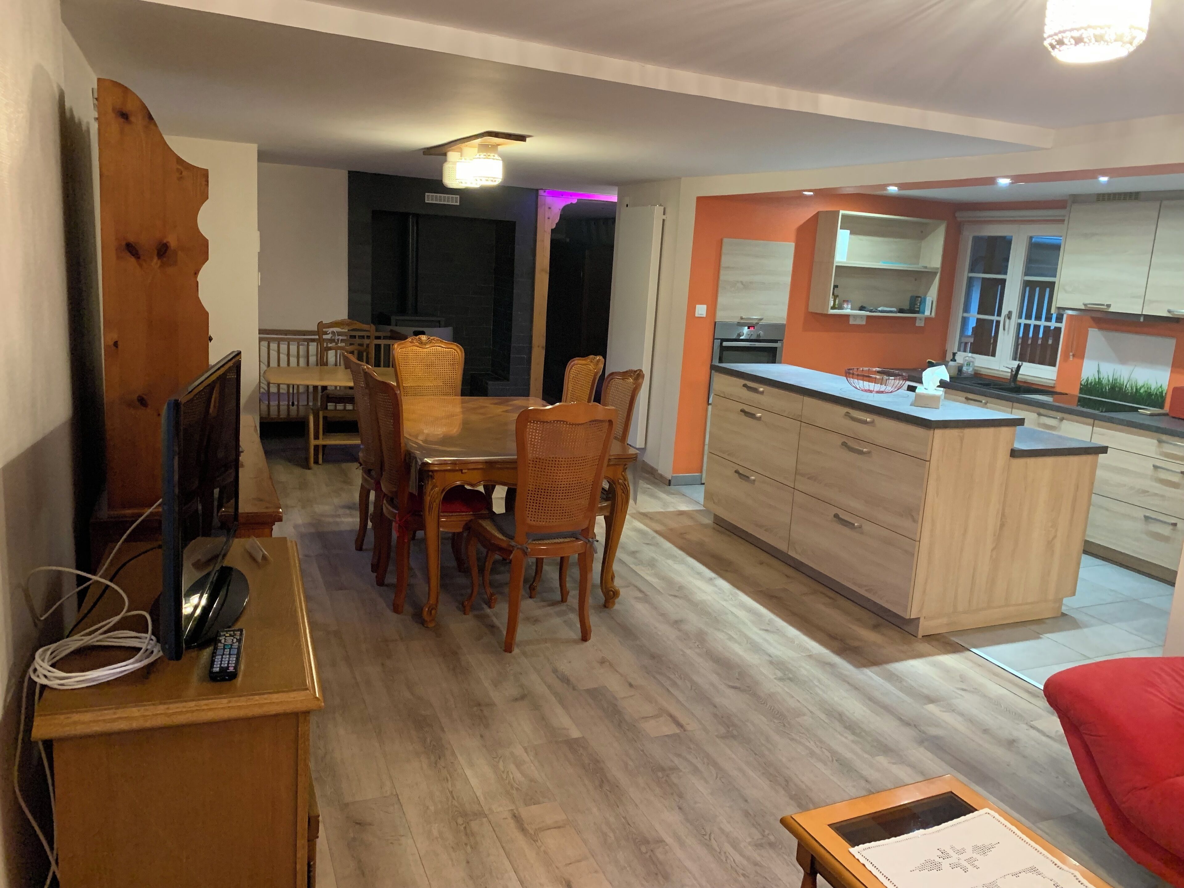 Eine moderne Küche mit Holzausstattung und einer zentralen Kochinsel, ein Essbereich mit Holztisch und Stühlen sowie ein Wohnbereich mit einem roten Sofa.
