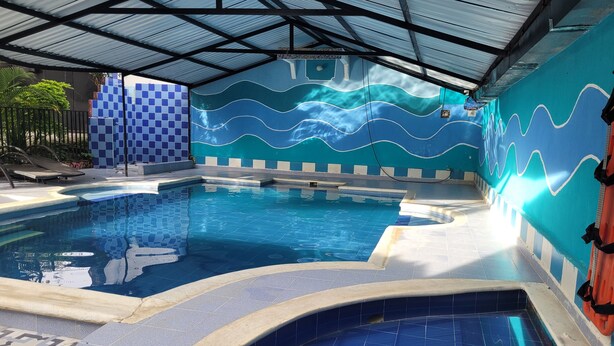 Ein Schwimmbecken mit einer wellenförmigen, blau-weißen Fliesenwand, einer gefliesten Liegefläche und einer Metall-Dachkonstruktion.