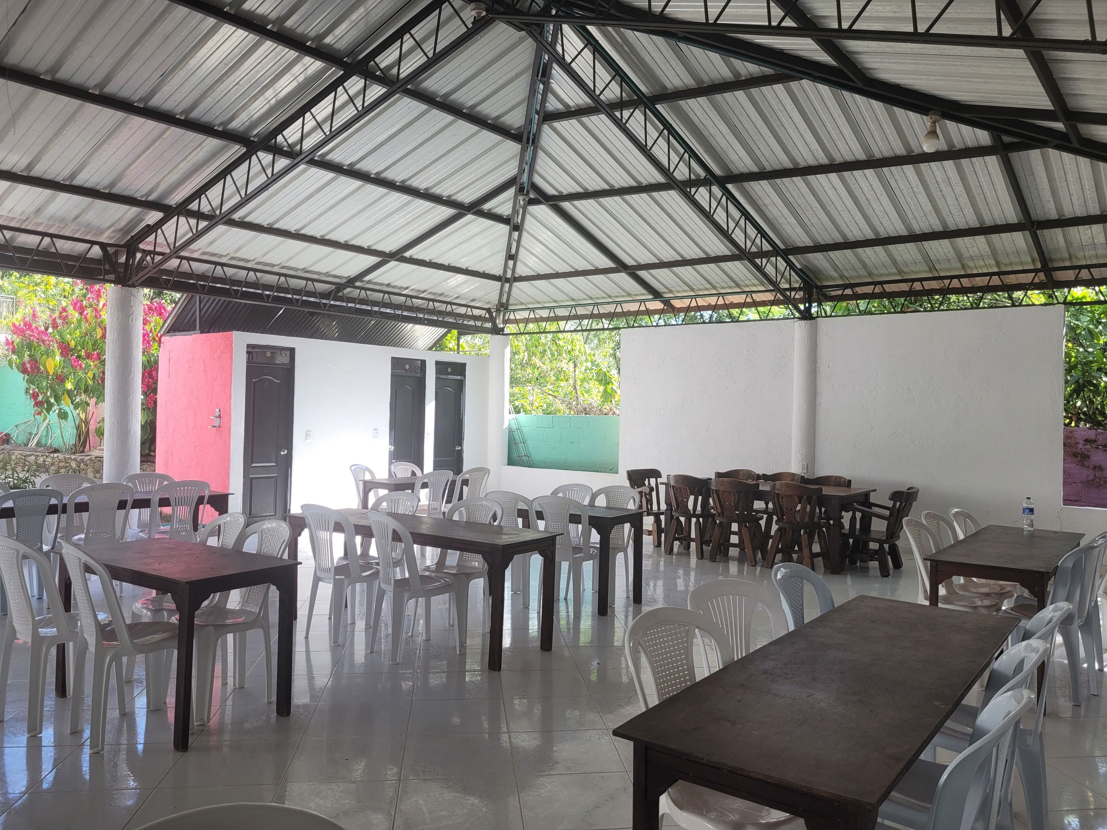 A covered outdoor dining area with tables and chairs, a tiled floor, and a metal roof structure.