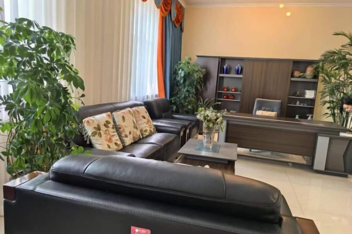 A modern living room with a black leather sofa, a coffee table, and a reception desk. There are potted plants and a bookshelf in the background.