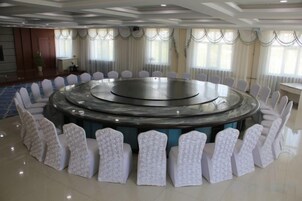 Un salón de conferencias con una mesa redonda rodeada de sillas, ventanas amplias con cortinas y suelo de baldosas.
