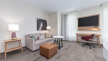 A hotel room with a sofa, ottoman, desk, chair, and a flat-screen TV mounted on the wall.