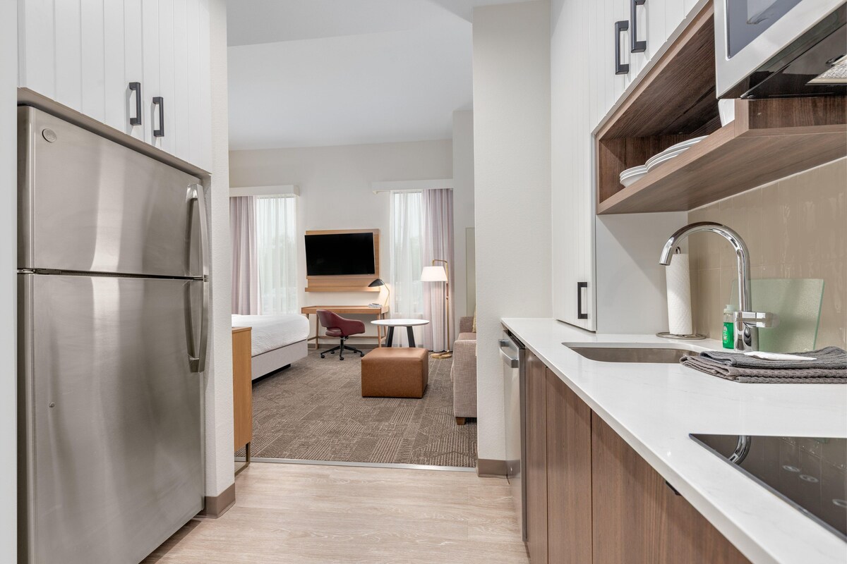 A modern hotel kitchen with stainless steel appliances, a dining area with a table and chairs, and a view into a bedroom with a bed and curtains.