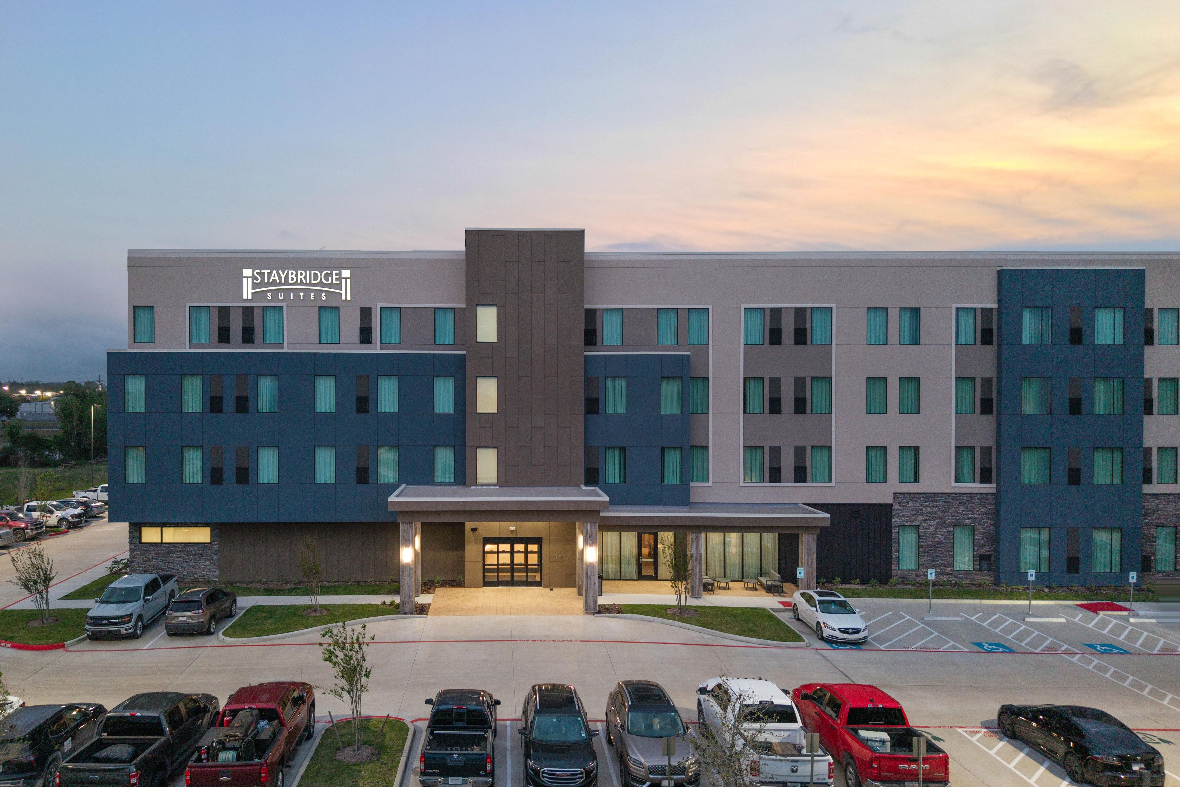 A Staybridge Suites hotel with a modern design, featuring multiple floors and a parking lot with various vehicles.