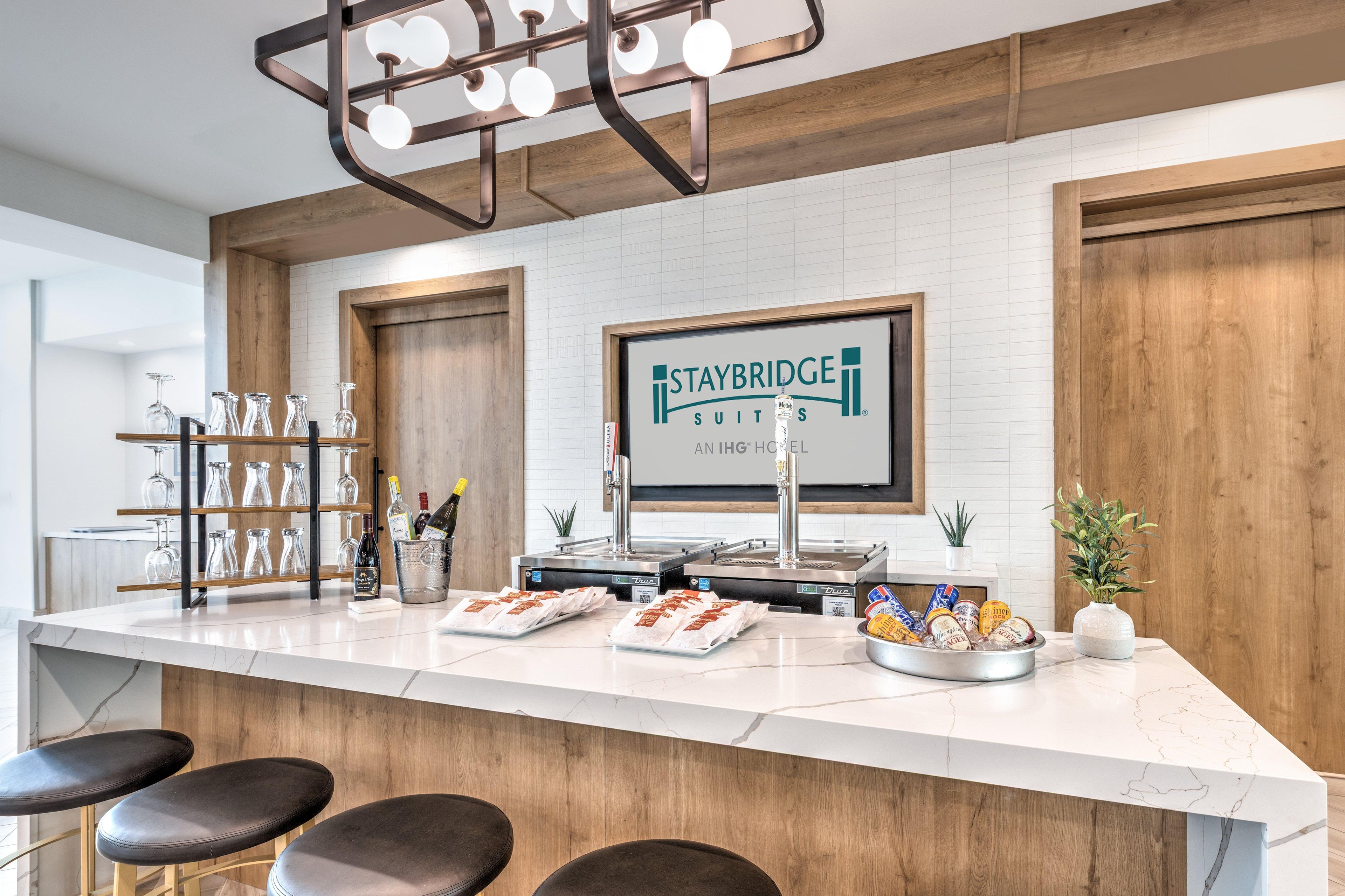 Angolo bar di un hotel con un bancone in marmo, sgabelli da bar, un cartello con la scritta Staybridge Suites e diversi snack e bevande.