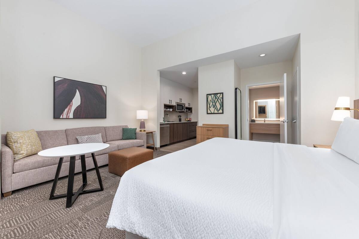 Habitación de hotel con una cama grande, un sofá, una mesita, una kitchenette y un baño visible a través de una puerta abierta.