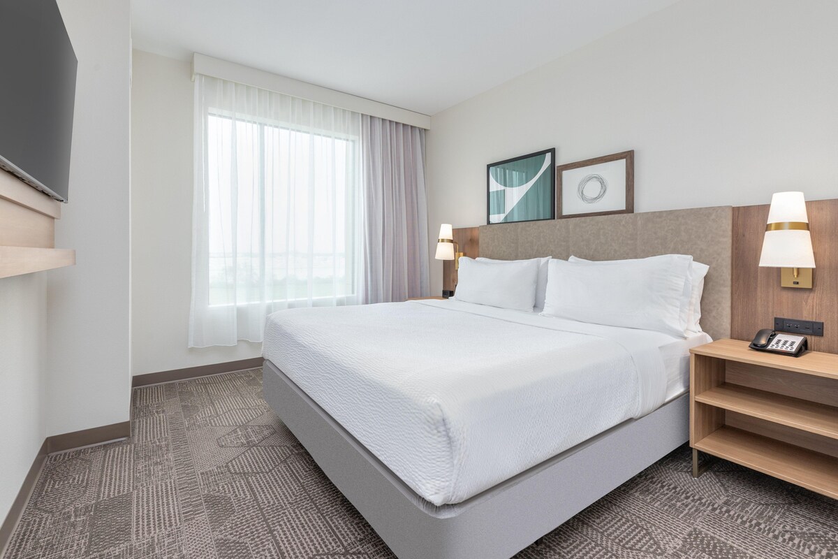 A hotel room with a large bed, two bedside tables with lamps, a flat-screen TV, and a window with curtains.