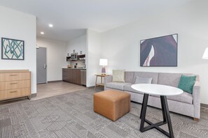 A modern living room with a grey sofa, a round white coffee table, and a brown ottoman. There is a kitchen in the background with a microwave and cabinets. - Staybridge Suites Port Arthur Mall Area (Port Arthur)