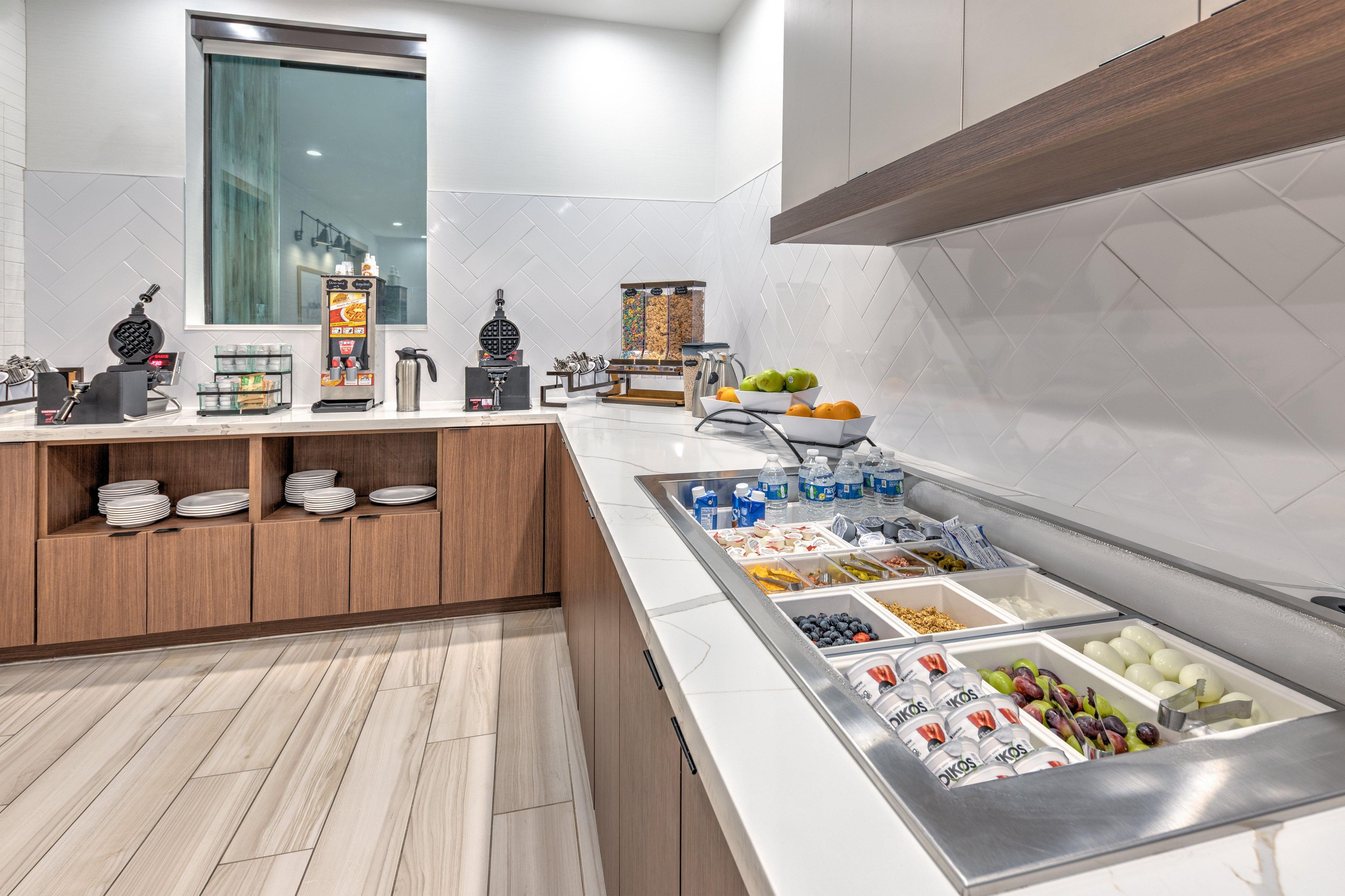 A modern kitchen with a breakfast bar, white tiles, wooden cabinets, and a variety of food and drink options.