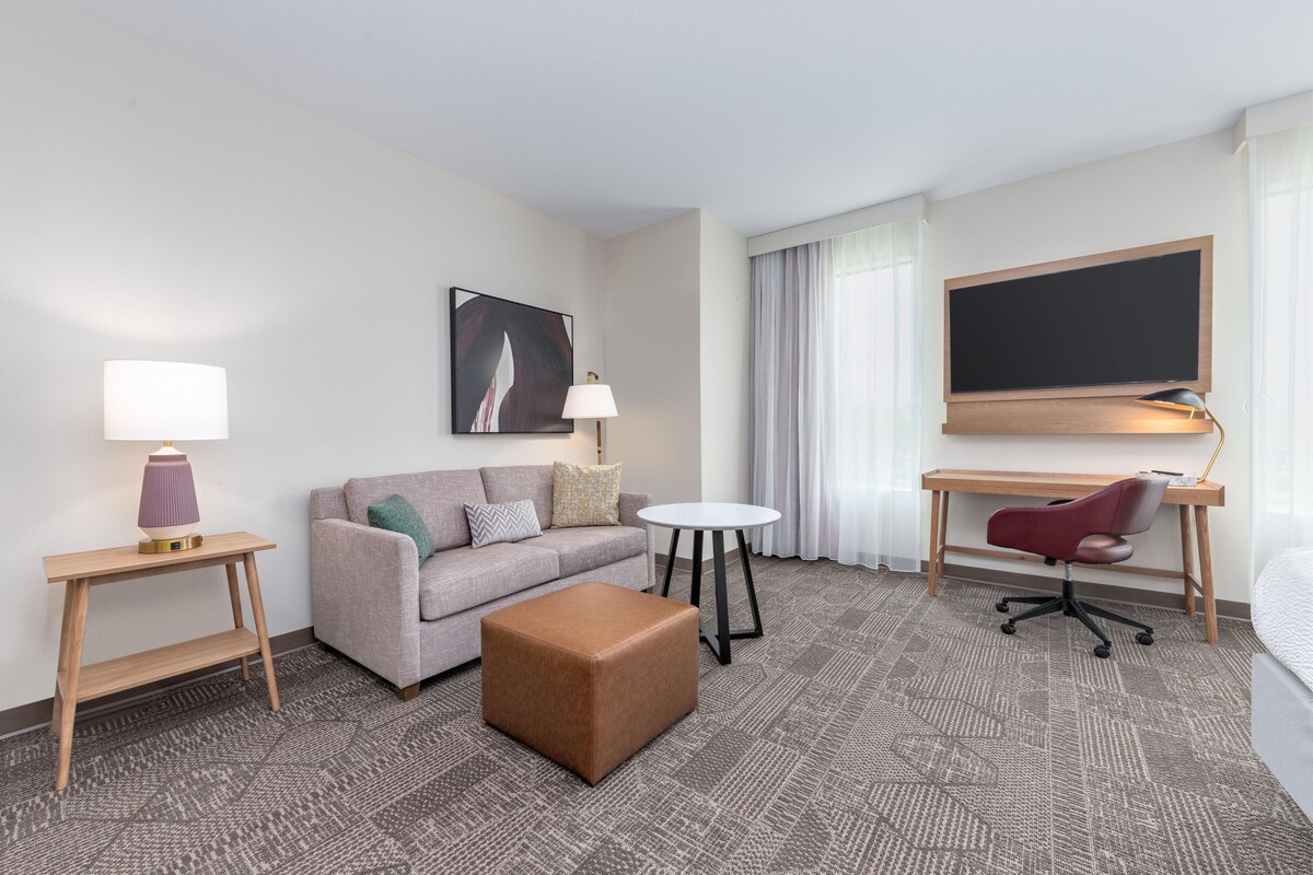 A hotel room with a sofa, ottoman, desk, chair, and a flat-screen TV mounted on the wall.