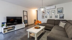En moderne stue med et fladskærms-tv, et hvidt sofabord med en vase blomster, en grå sofa og en vægmonteret aircondition.