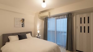 A hotel room with a large bed, a bedside lamp, an air conditioning unit, a window with curtains, and a wardrobe. - Dotonbori East Residence Haru (Osaka)