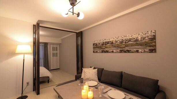 A modern living room with a dining table set for two, a sofa, a floor lamp, and a painting on the wall. - Dotonbori East Residence Haru (Osaka)