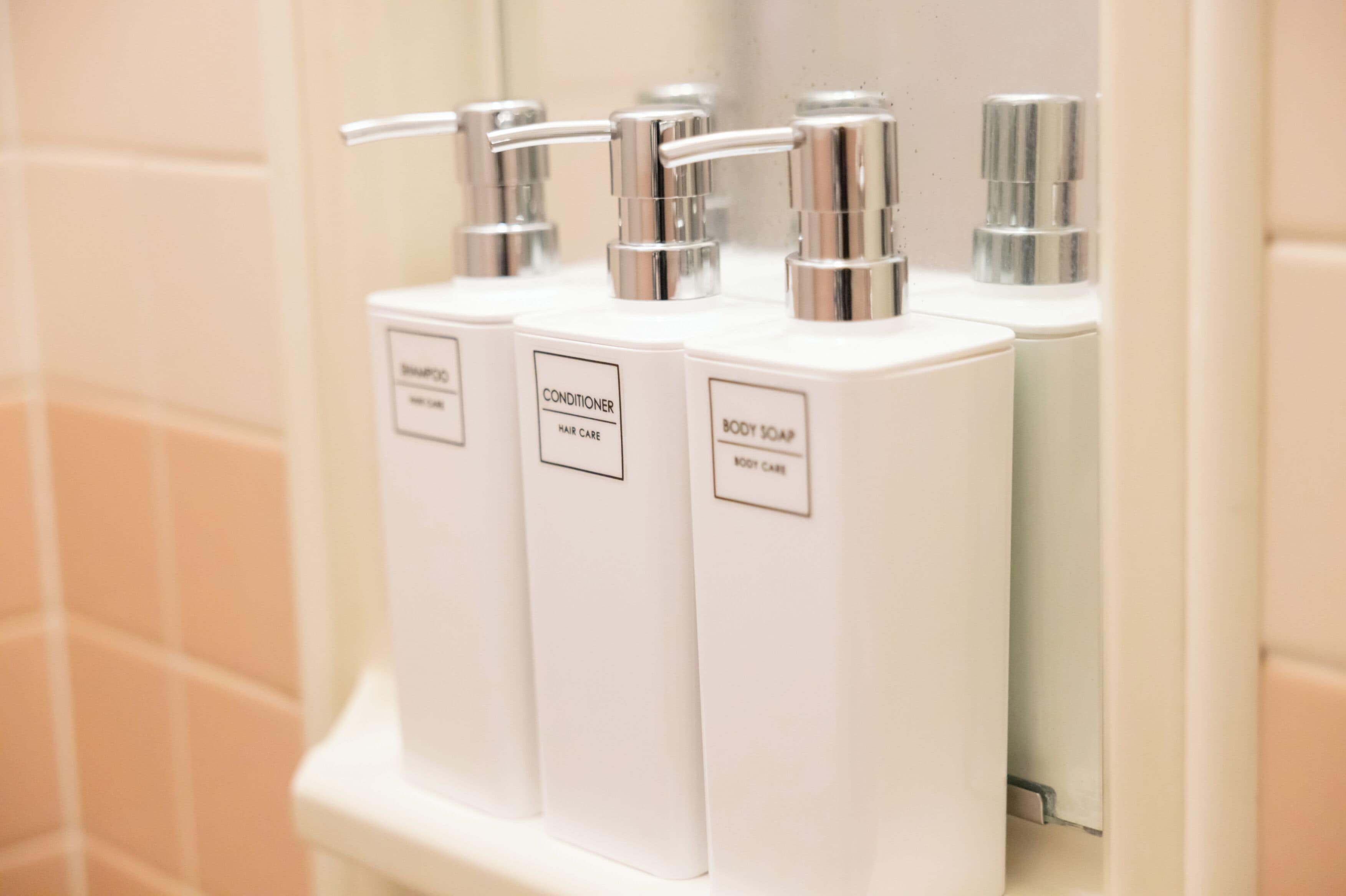 A bathroom with three white dispensers labeled for shampoo, conditioner, and body soap.