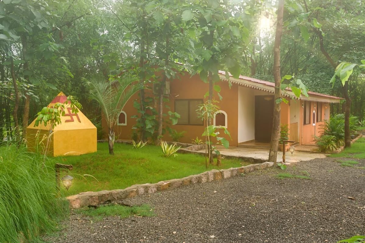A small, single-story building with a tiled roof and a porch, surrounded by lush greenery and a gravel path.